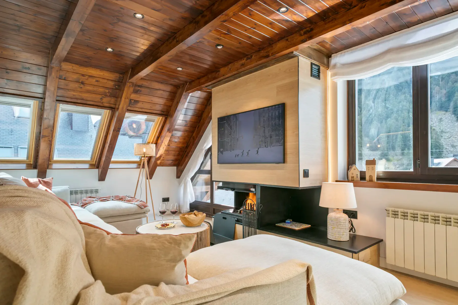 Modern living room with light wood ceiling beams, large windows, a gray sectional sofa, a wooden coffee table, poufs, and a fireplace with a TV mounted next to it. Scenic mountains are visible outside.