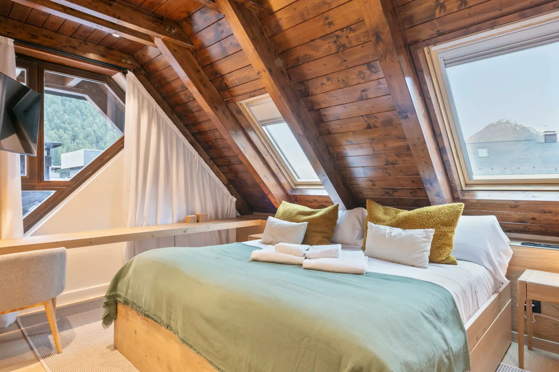 A cozy attic bedroom with wooden walls and ceiling, a bed with folded towels, a window with a scenic mountain view, and an adjacent bathroom visible through a doorway.