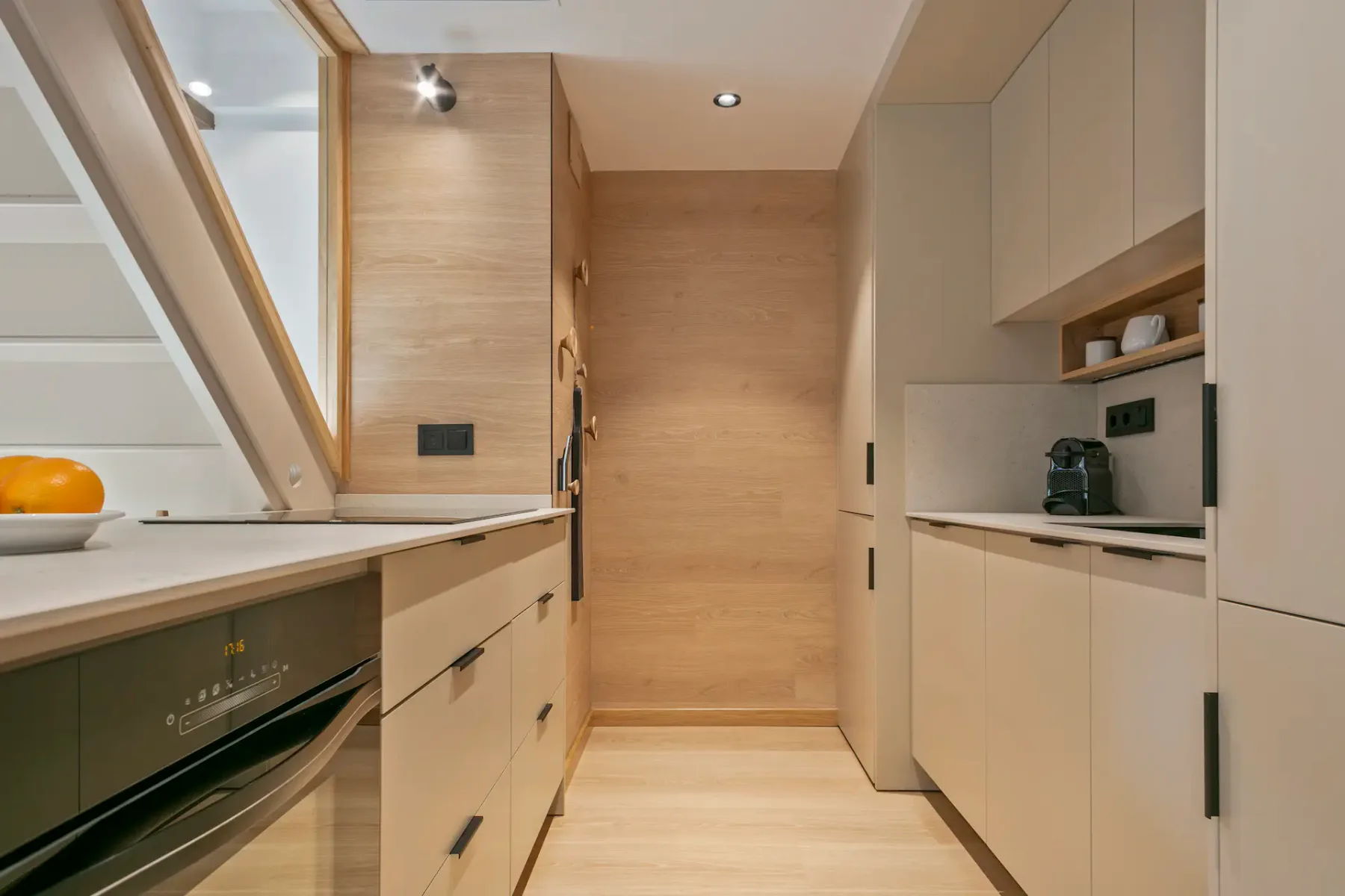 A narrow kitchen with light cabinetry, built-in appliances, and wood-paneled walls.