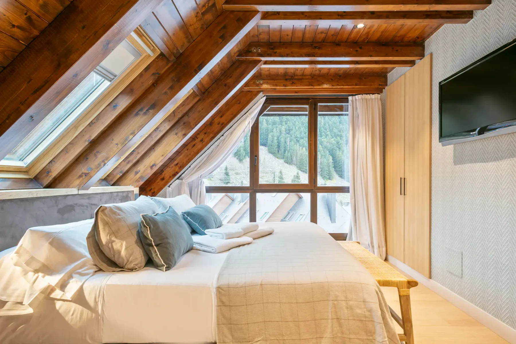 An attic bedroom with a bed beneath sloped wooden beams and windows.
