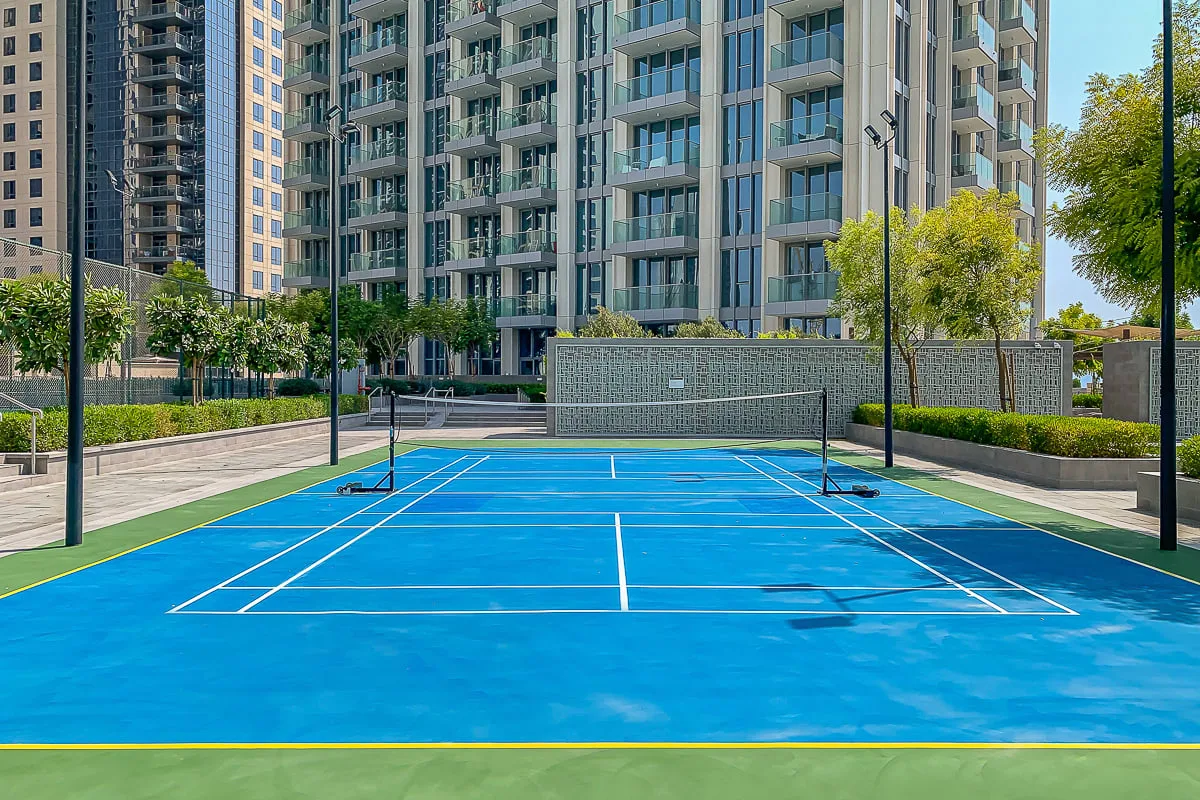 Una cancha de tenis azul al aire libre está junto a senderos ajardinados y edificios altos.