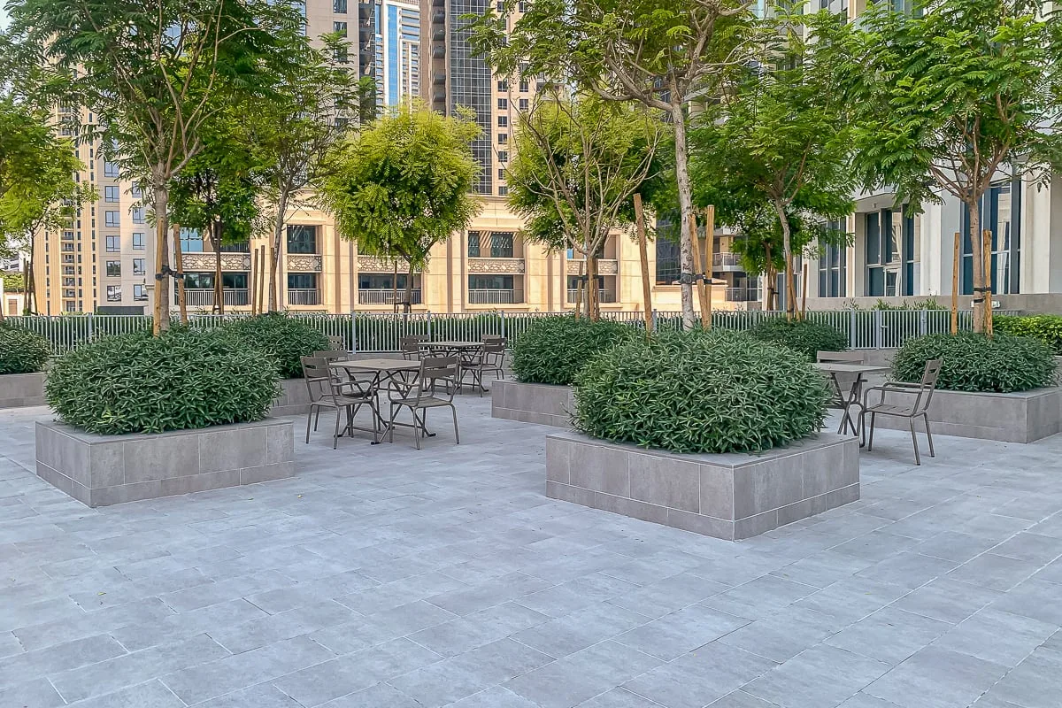 Una zona de estar al aire libre tiene mesas y sillas entre jardineras elevadas y árboles.