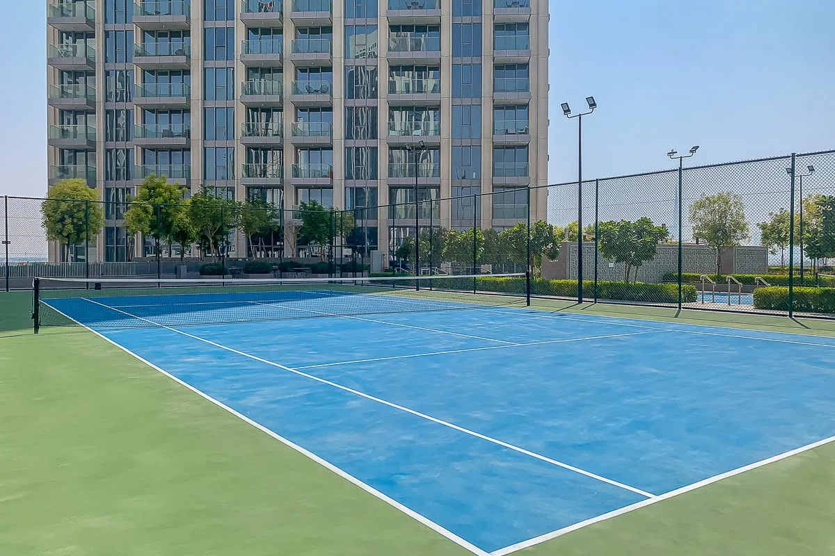 Una pista de tenis azul está junto a un edificio alto de apartamentos y un recinto cercado.