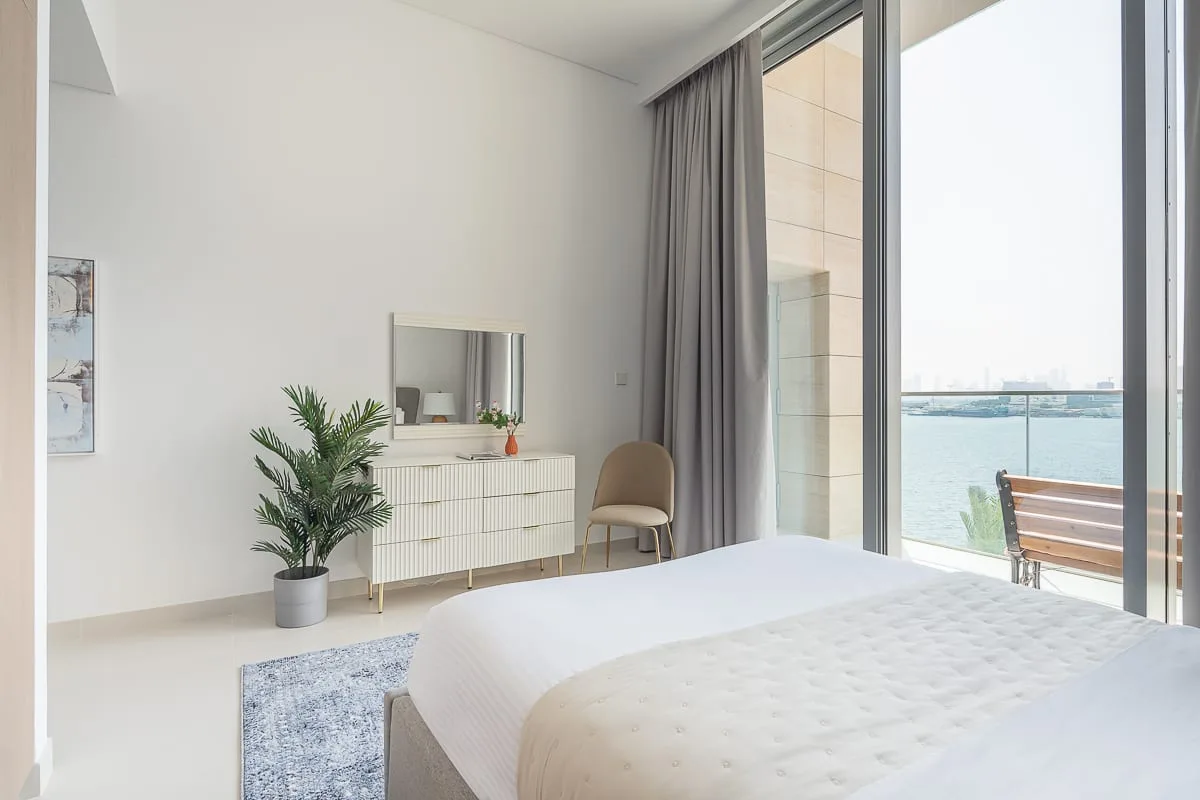 Un dormitorio luminoso con una cama blanca, una cómoda y puertas corredizas de vidrio.