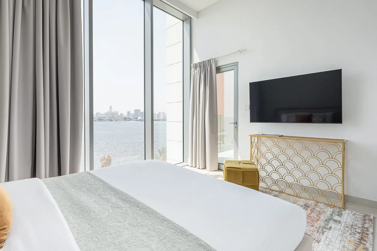 Un dormitorio luminoso con una cama, un televisor en la pared y grandes ventanas con vistas al agua.