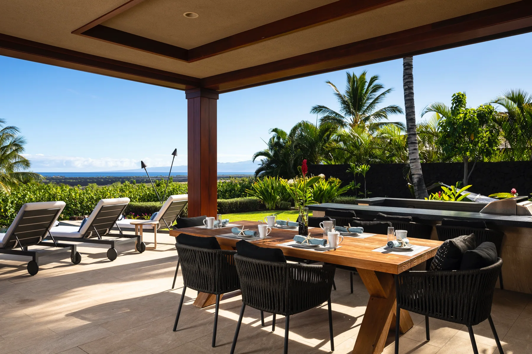 An outdoor dining area under a covered patio overlooks a garden and the ocean.