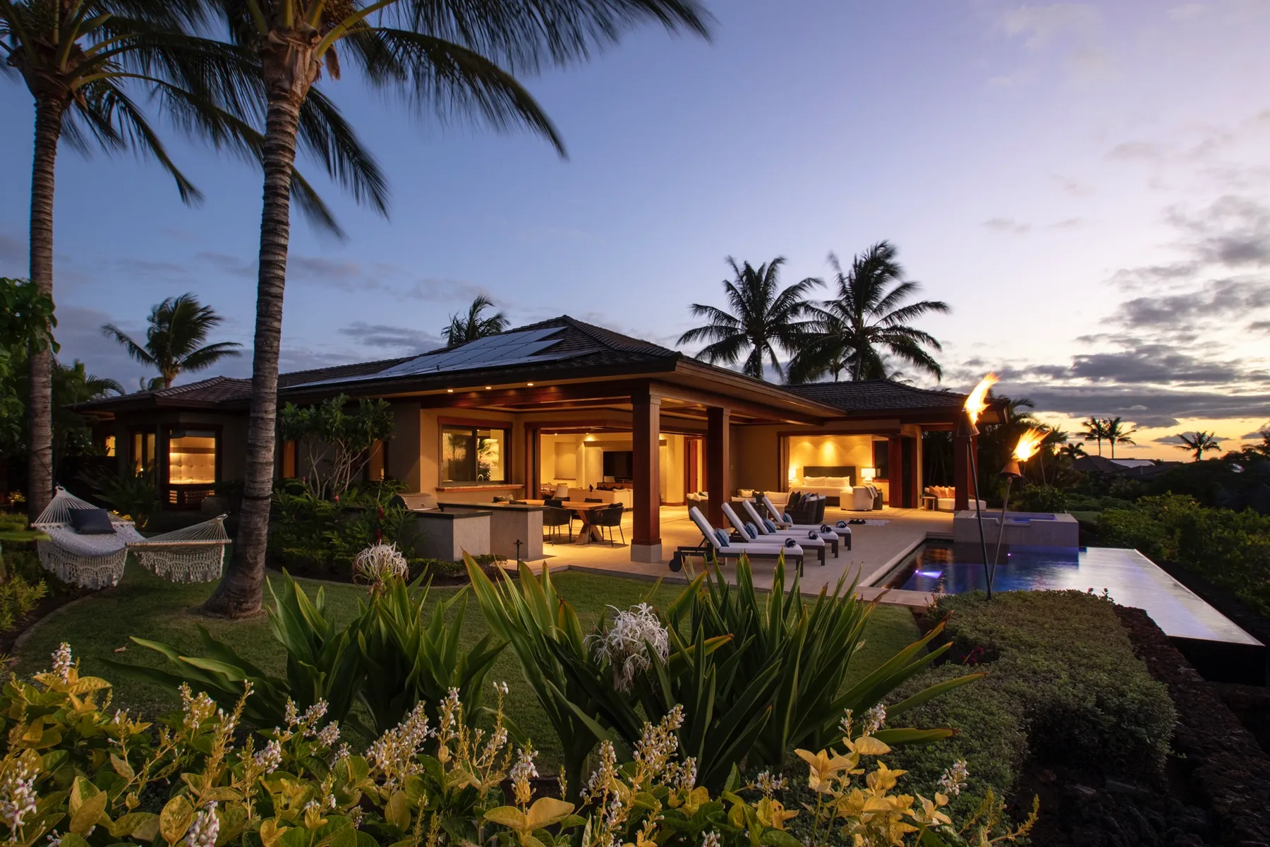 A tropical resort building glows at dusk beside a swimming pool and palm trees.