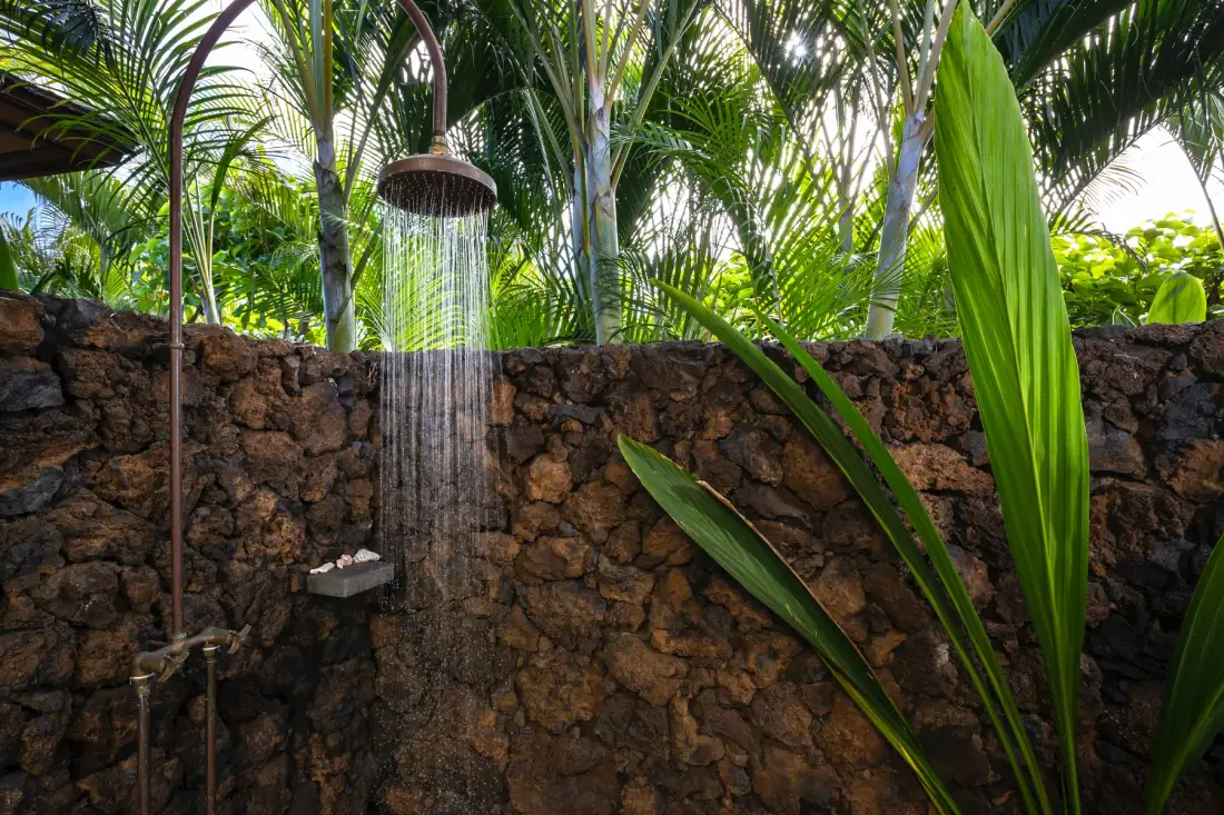 An outdoor shower sprays water beside a stone wall, surrounded by tropical plants.