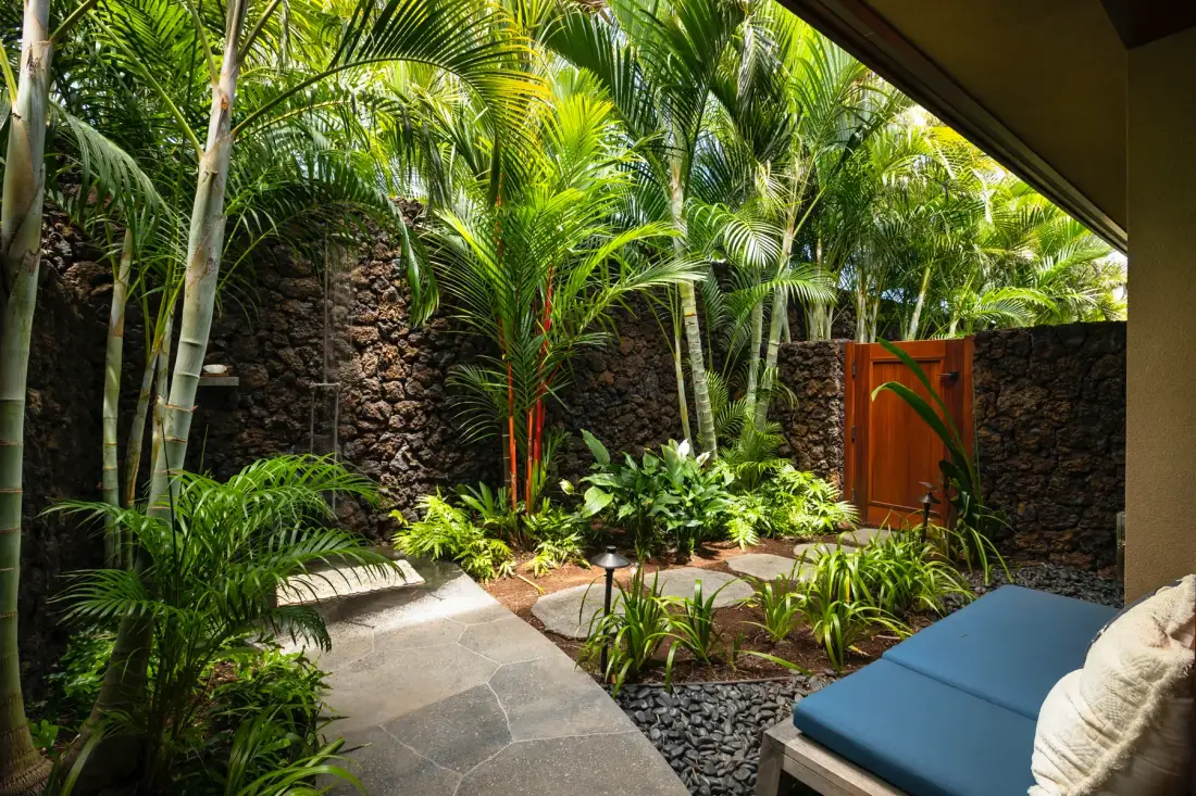 A lush tropical courtyard with stone walls, leafy plants, and a blue lounge seat.