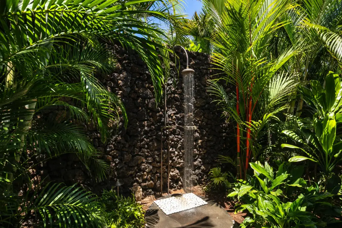 An outdoor shower stands against a stone wall amid dense tropical plants.