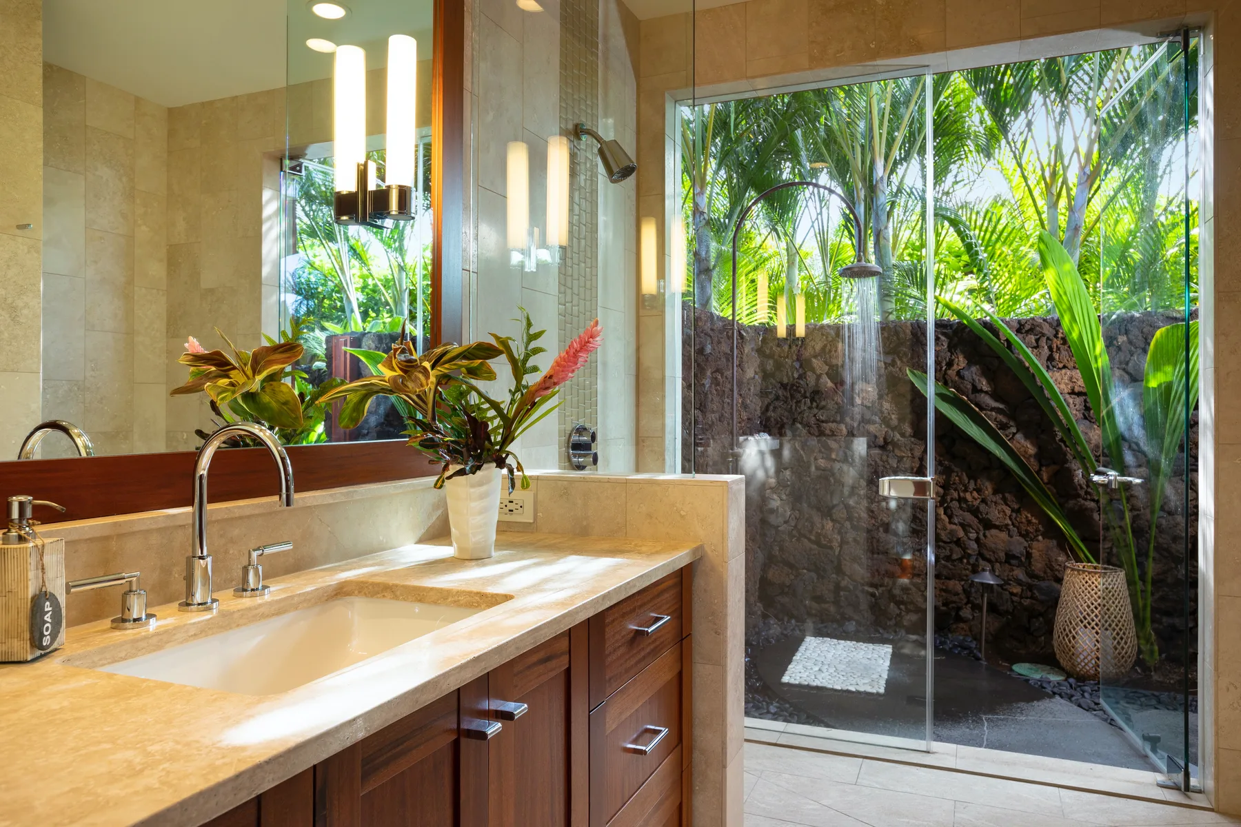 A bathroom vanity with a wide mirror beside a glass shower overlooking greenery.