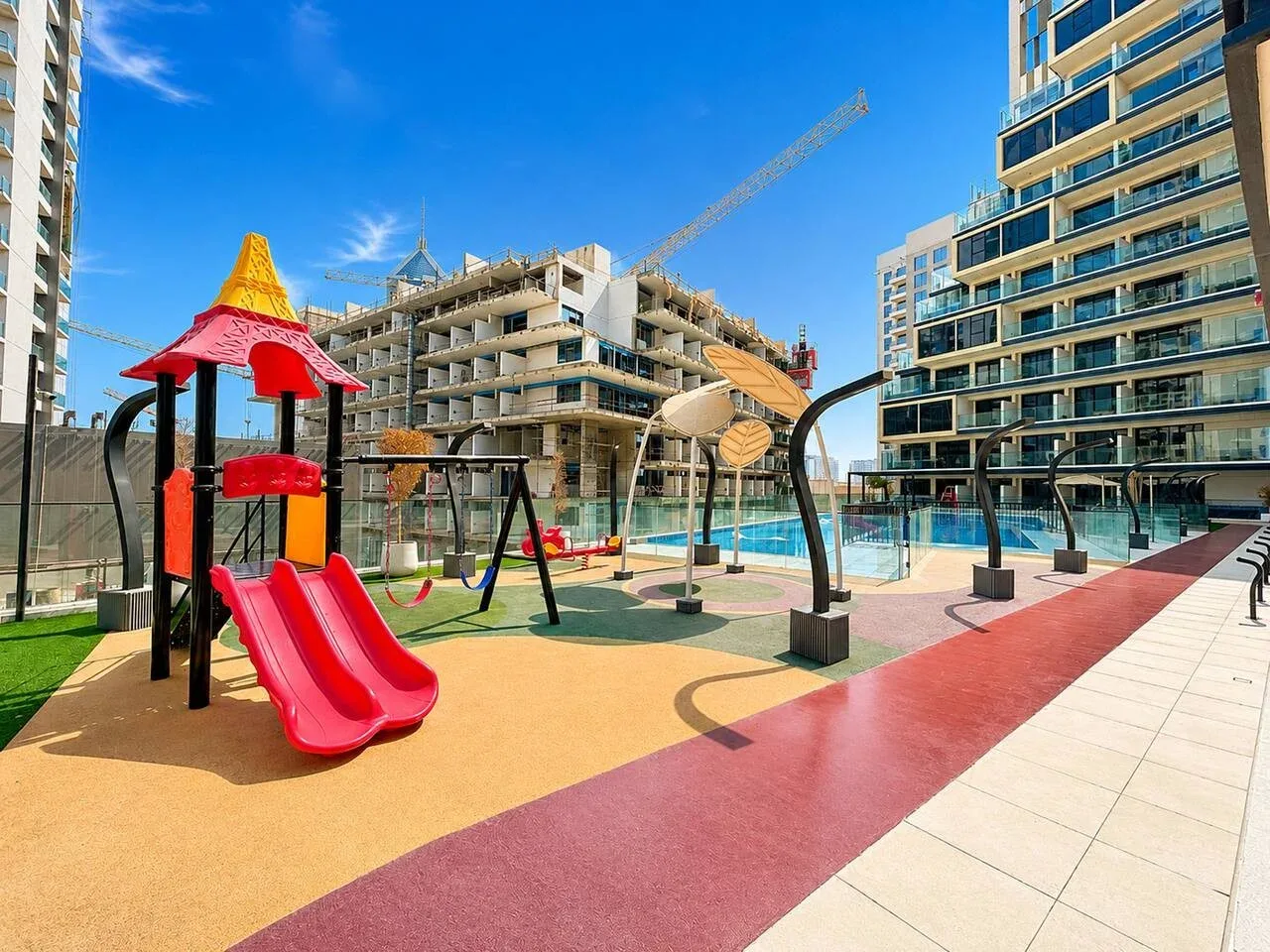 Un parque infantil al aire libre con un tobogán rojo y columpios junto a una piscina.