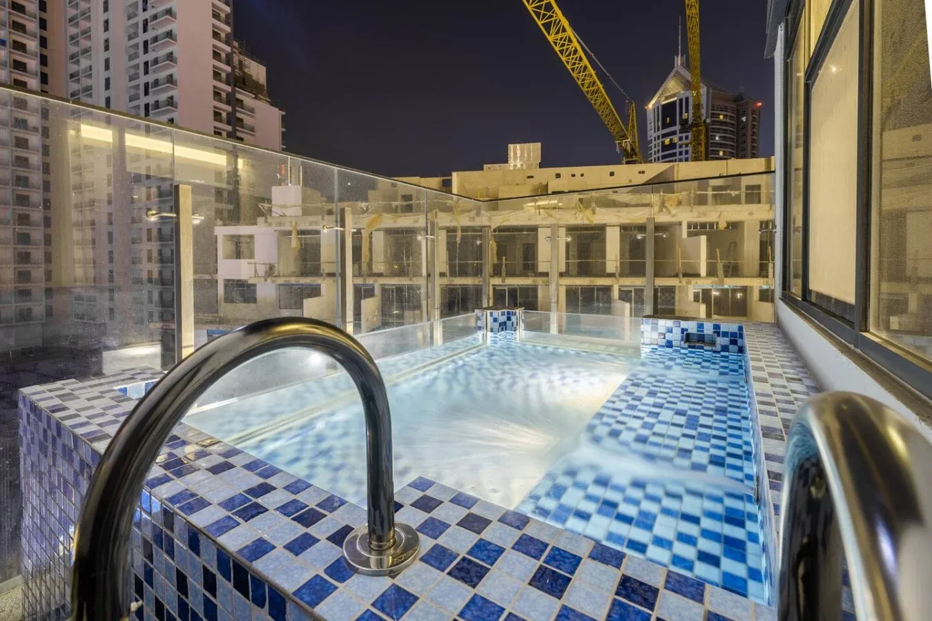Una piscina exterior al anochecer, rodeada de barandillas de vidrio y edificios.