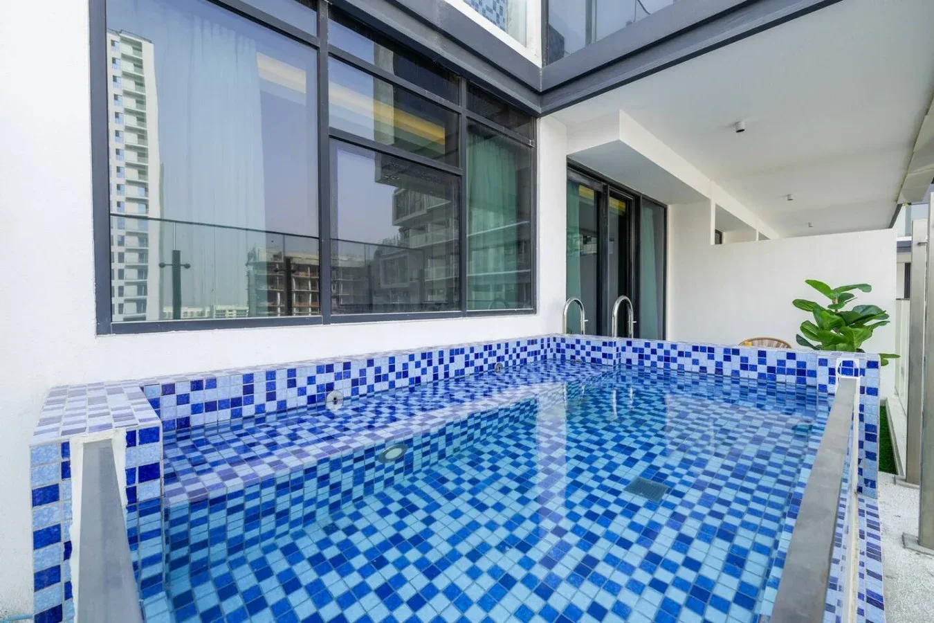 Una pequeña piscina de azulejos está junto a grandes ventanales en un patio moderno.