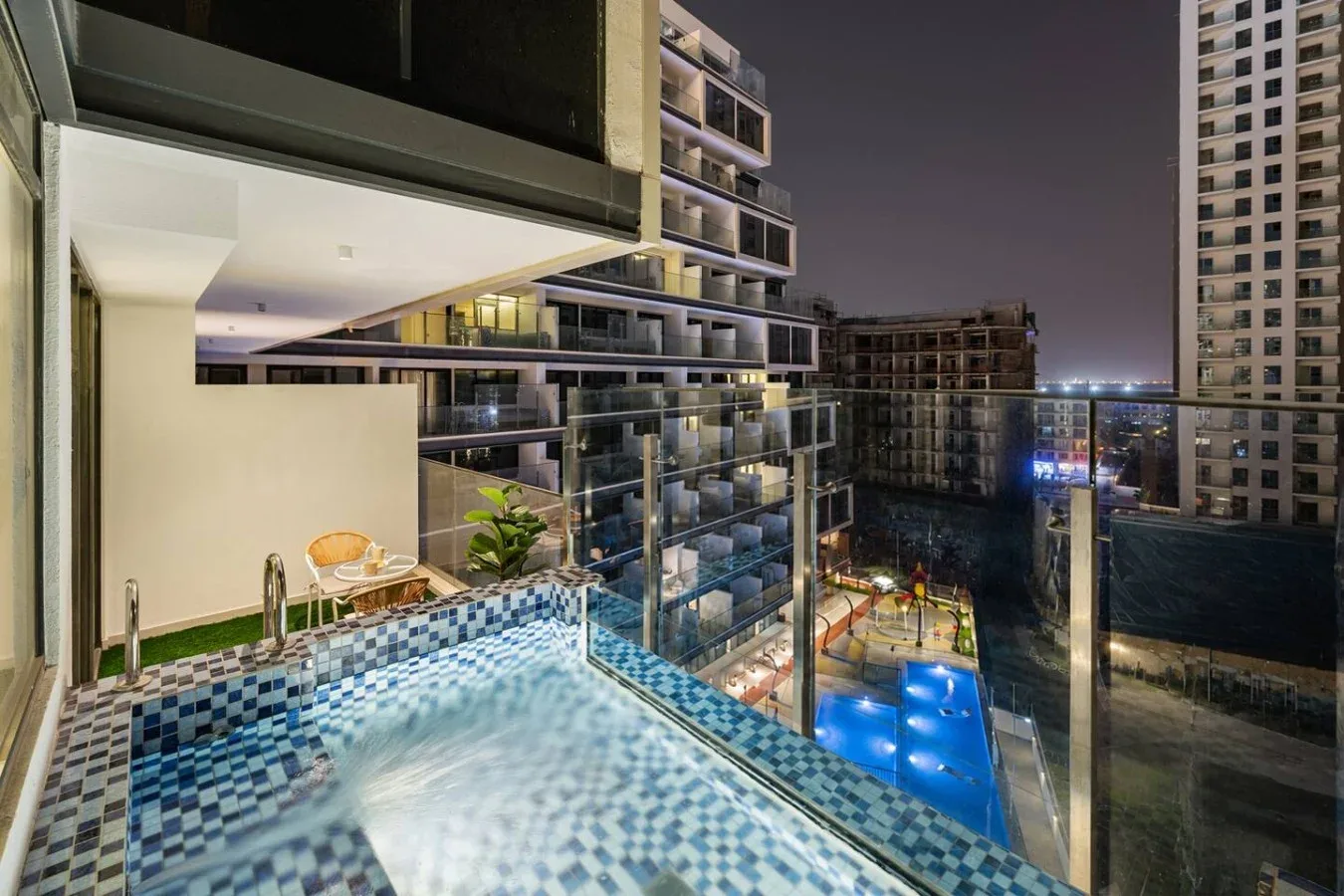 Una piscina exterior de azulejos tiene vistas a edificios altos iluminados por la noche.