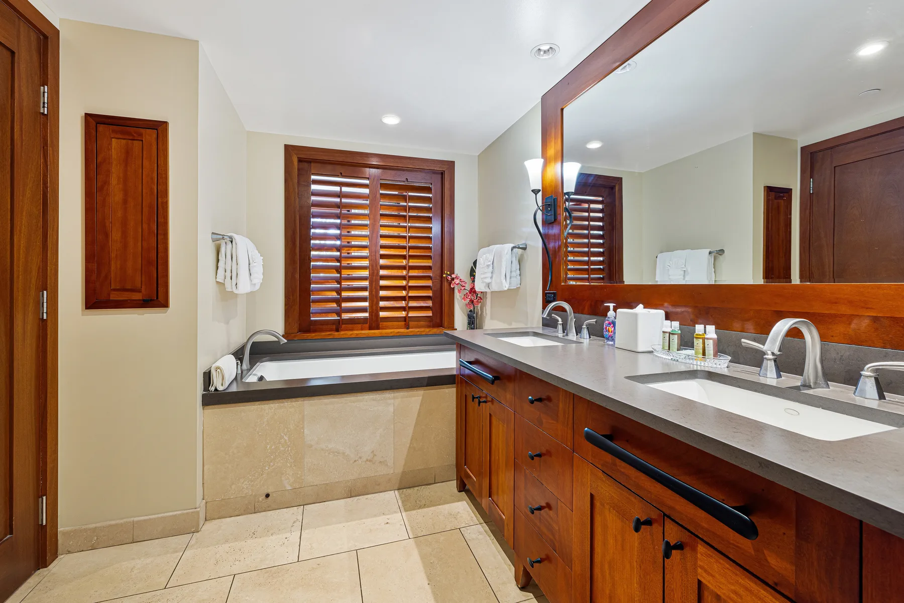 Un baño con un mueble largo, lavabos y una ventana con persianas de madera.