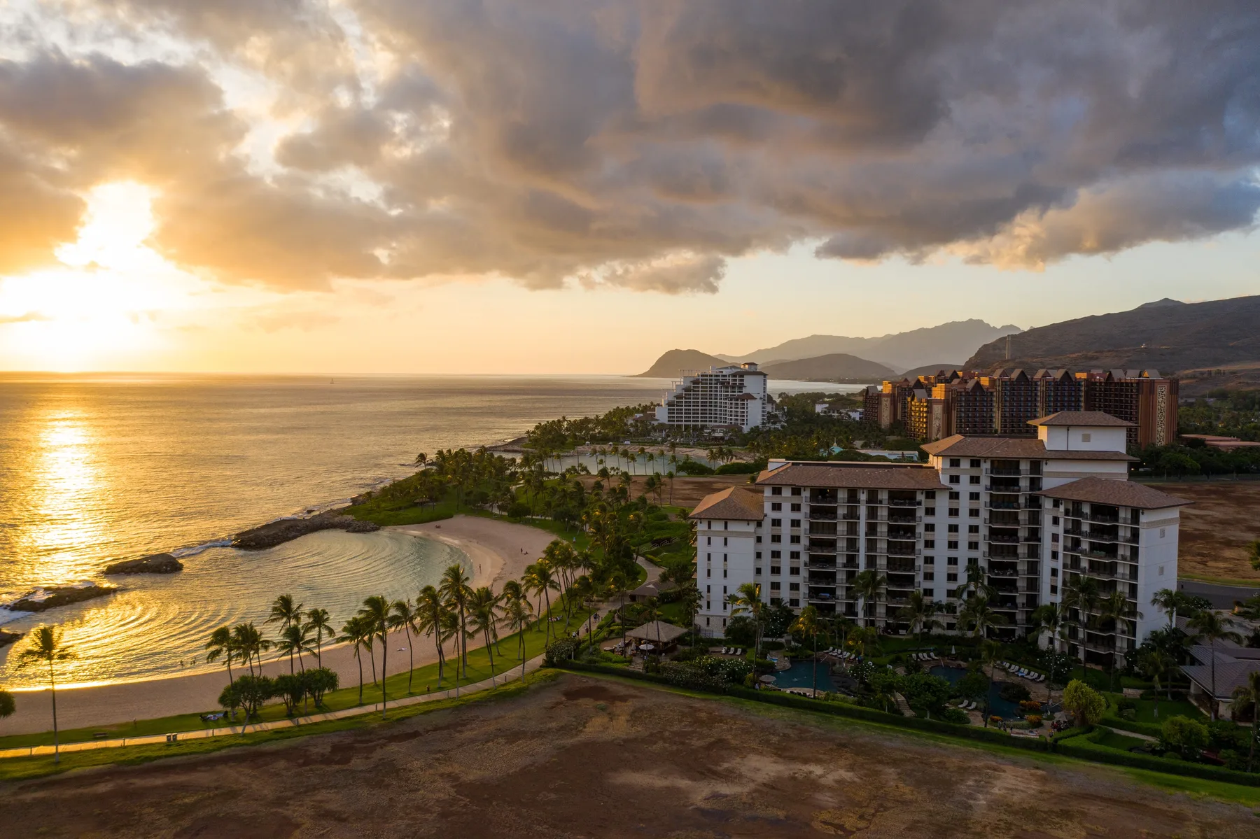 Edificios de un complejo costero dan a una playa bordeada de palmeras y al océano al atardecer.