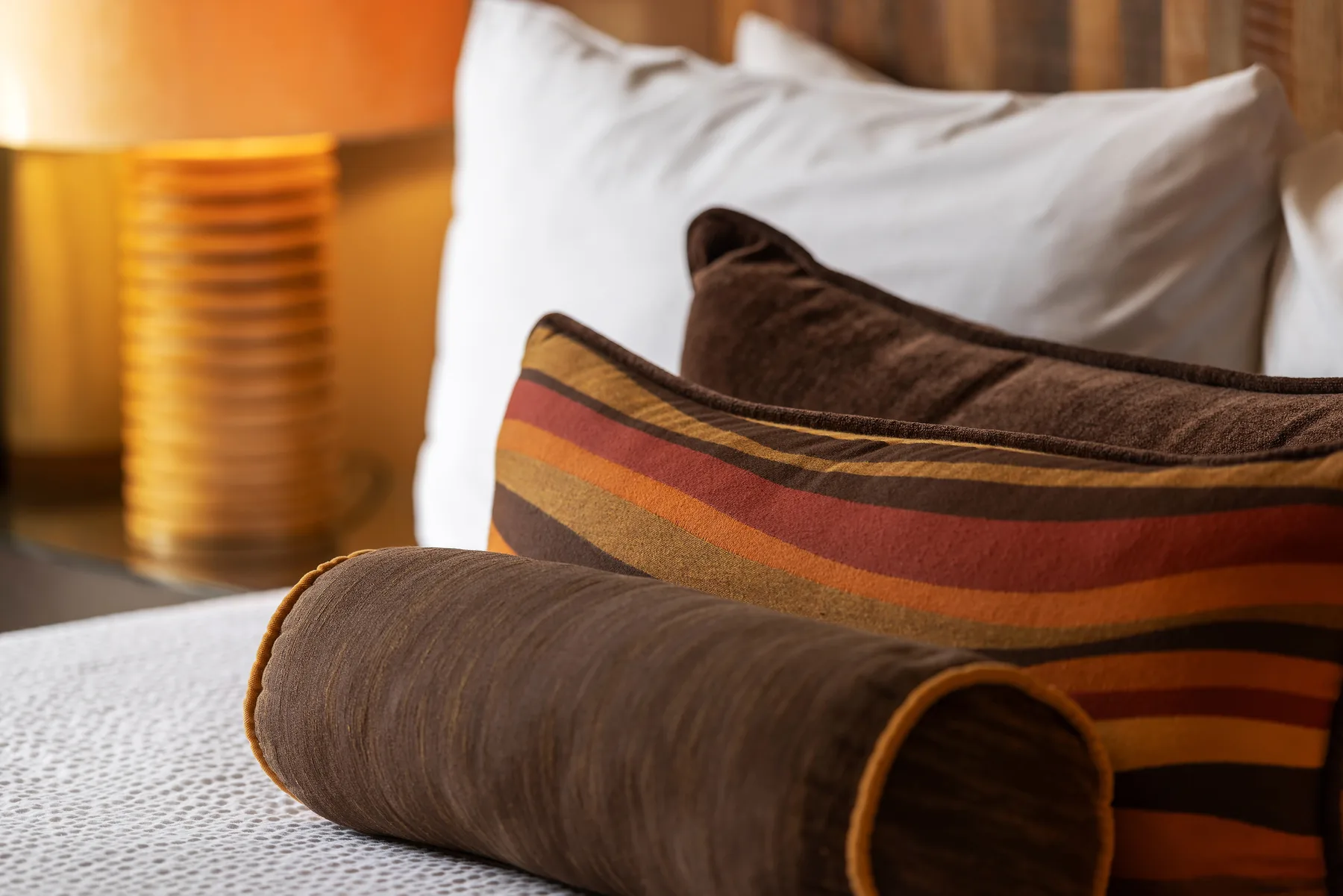 Una cama con almohadas superpuestas y un cojín cilíndrico marrón.