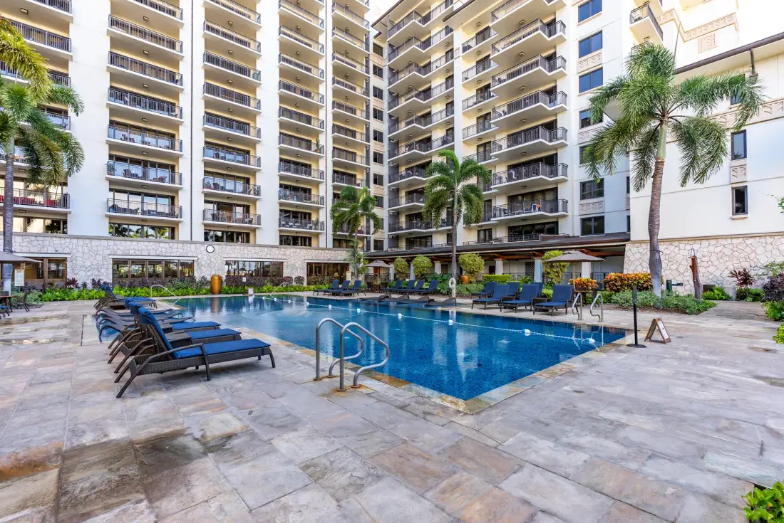 Una piscina exterior está rodeada de asientos y un edificio alto de apartamentos.