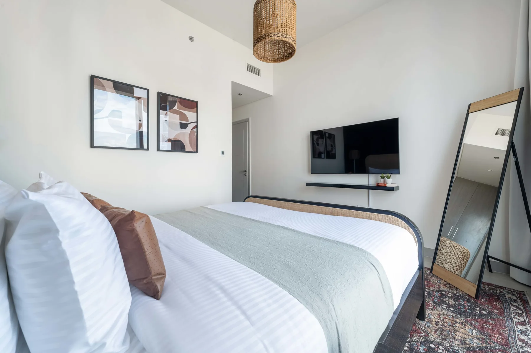 Un dormitorio luminoso con una cama bien tendida, un televisor en la pared y un espejo de cuerpo entero.
