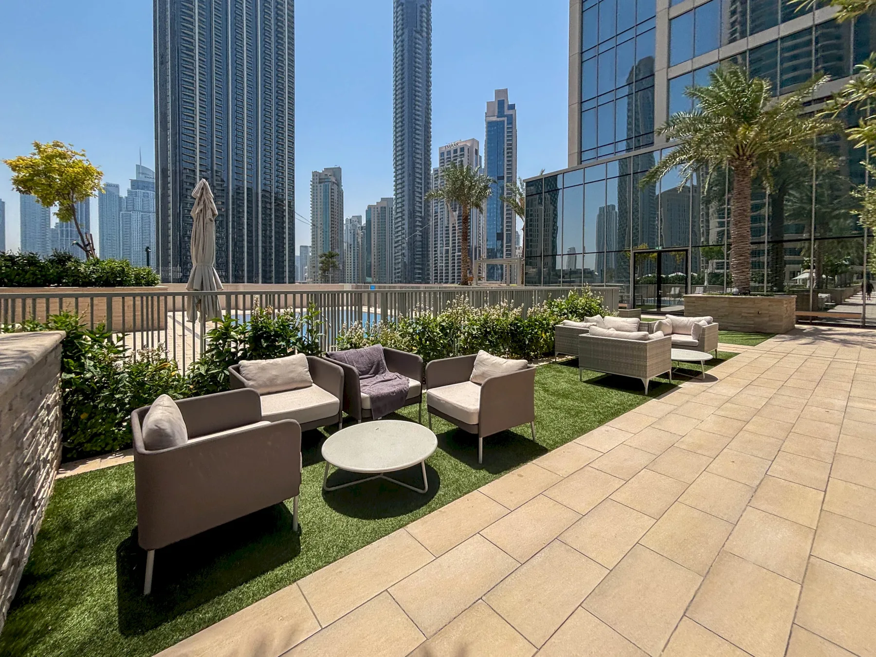 Una terraza al aire libre tiene asientos sobre césped junto a un camino pavimentado y edificios de vidrio.
