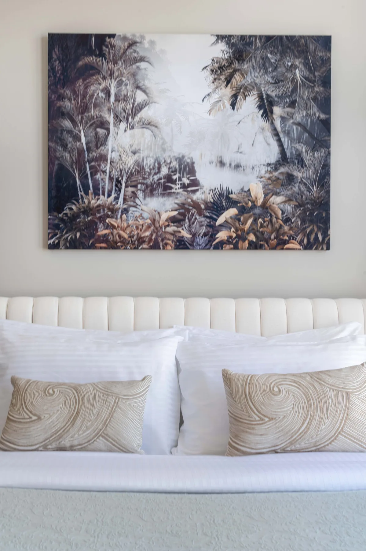 Una cama bien arreglada con cojines decorativos está debajo de una lámina enmarcada de un paisaje tropical.