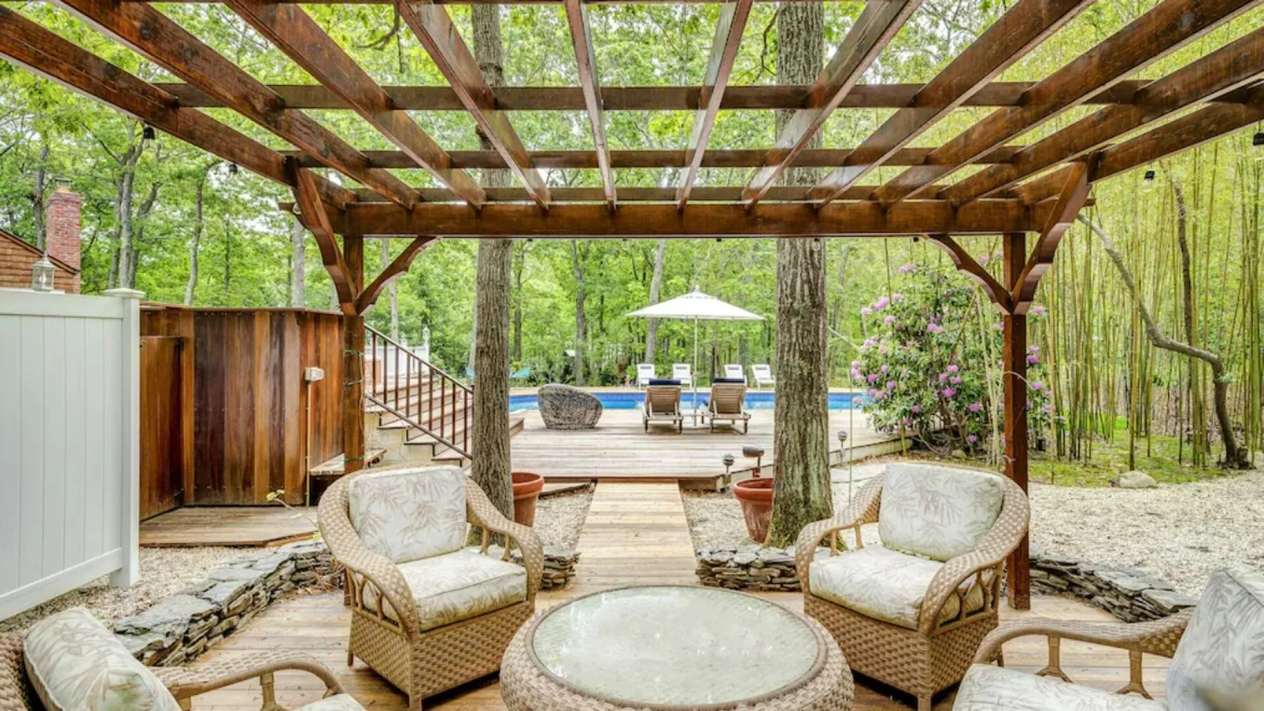 Una zona de estar exterior cubierta, con sillones acolchados frente a una piscina y un jardín.