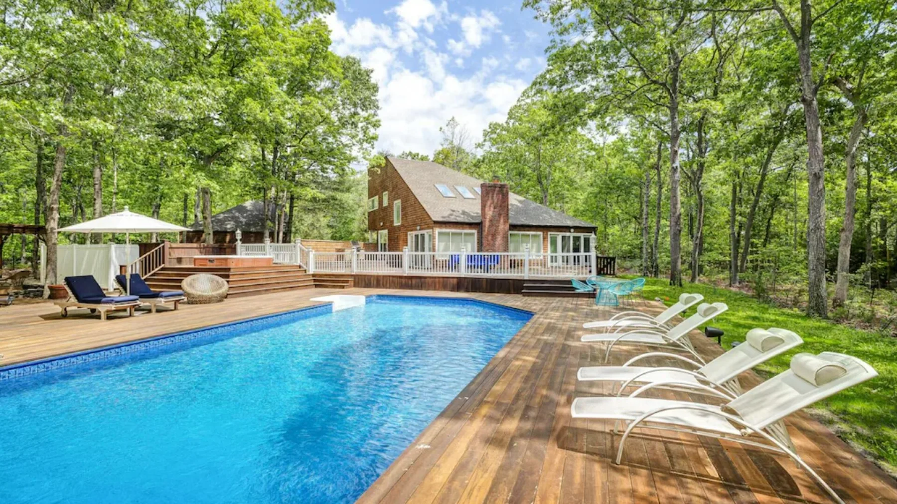 Una piscina al aire libre junto a una terraza de madera, asientos y una casa.