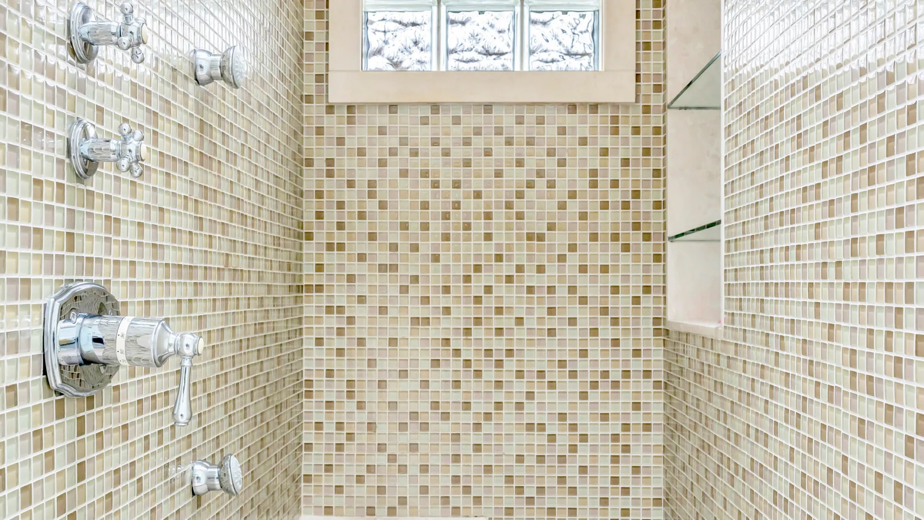 Una zona de ducha con azulejos, varios controles cromados y una ventana pequeña y alta.