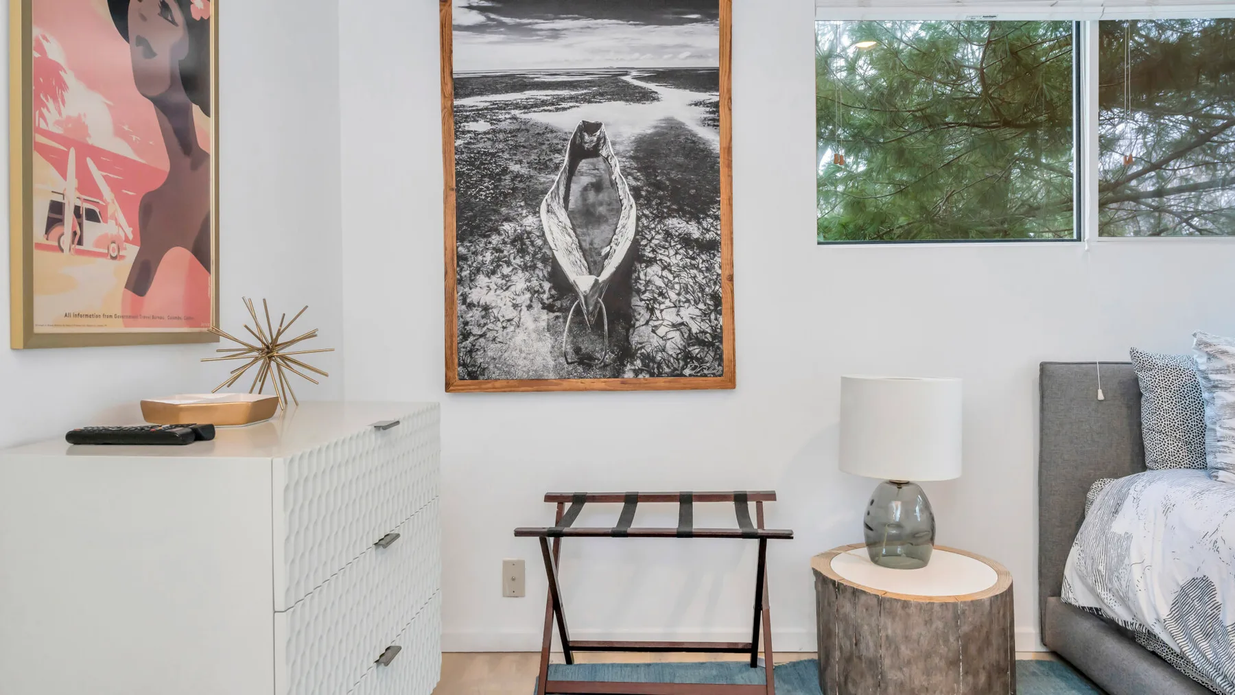 Un dormitorio luminoso con cuadros enmarcados, una cama y una ventana con vistas a los árboles.