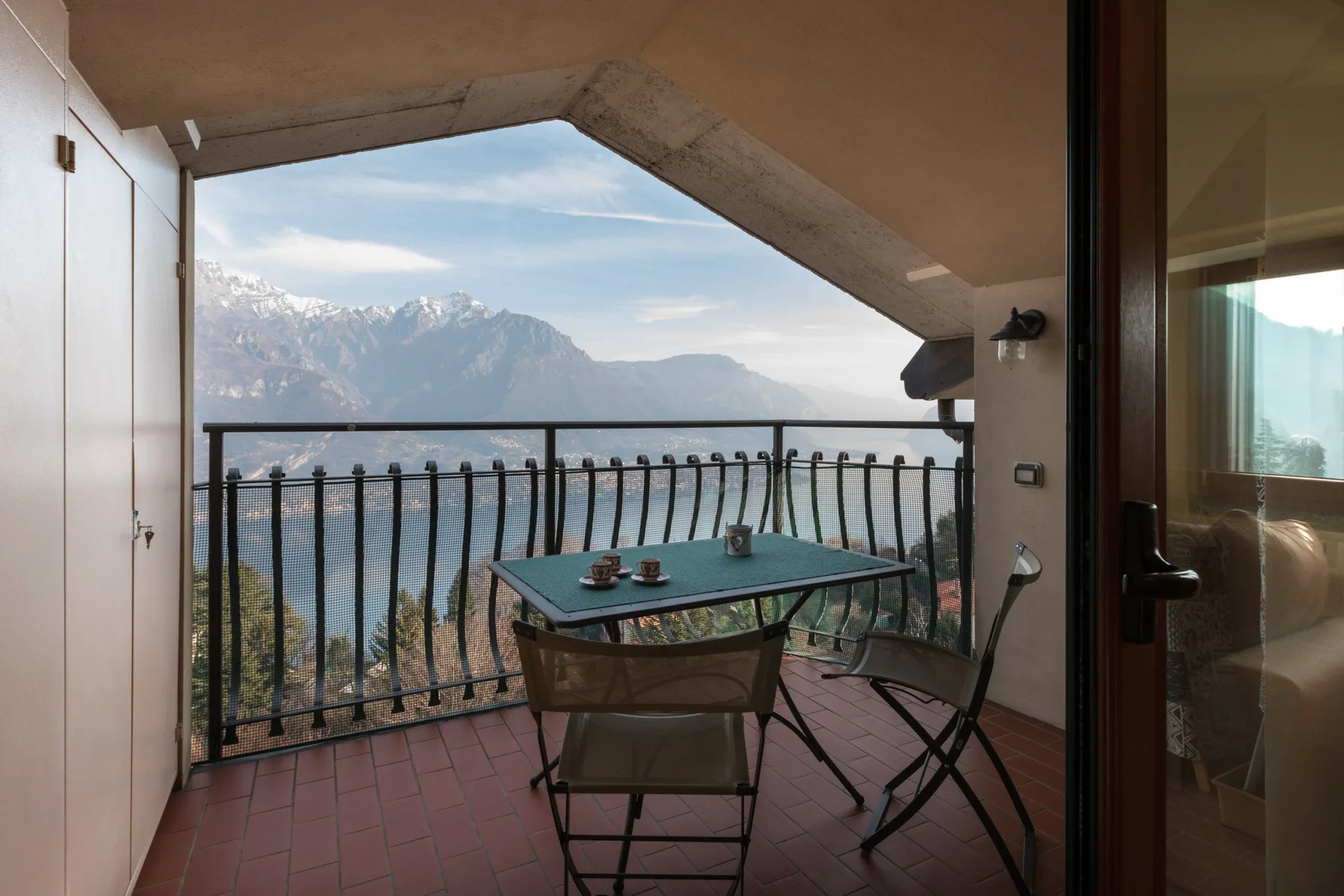 A covered balcony with a table and seating overlooks a lake and mountains.