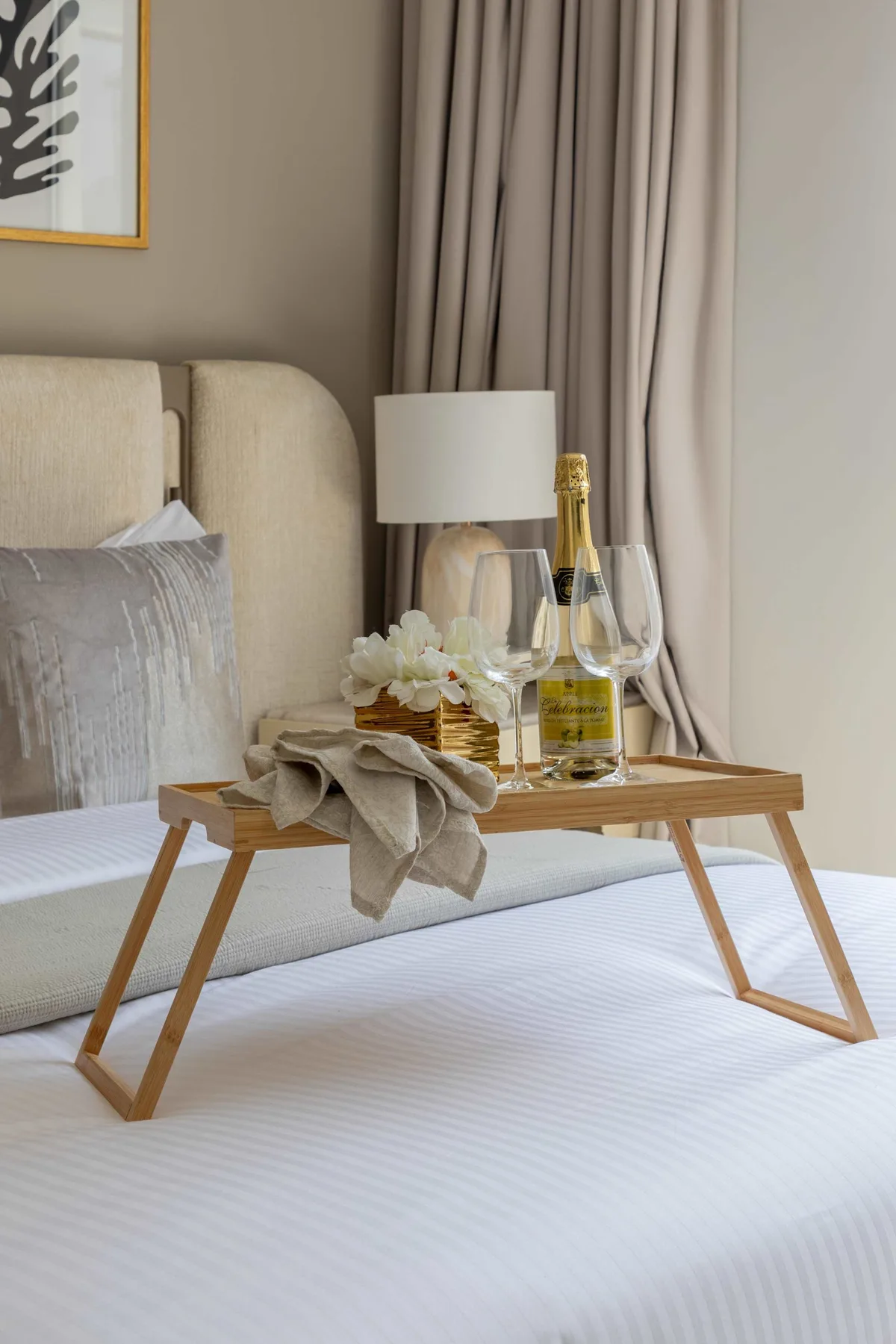 A neatly made bed with a wooden tray holding flowers and a bottle.
