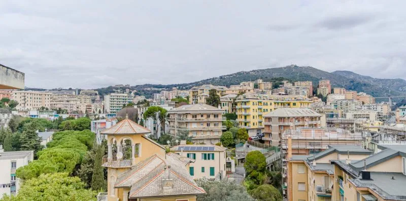 A cityscape of closely spaced buildings and green hills beneath a cloudy sky.