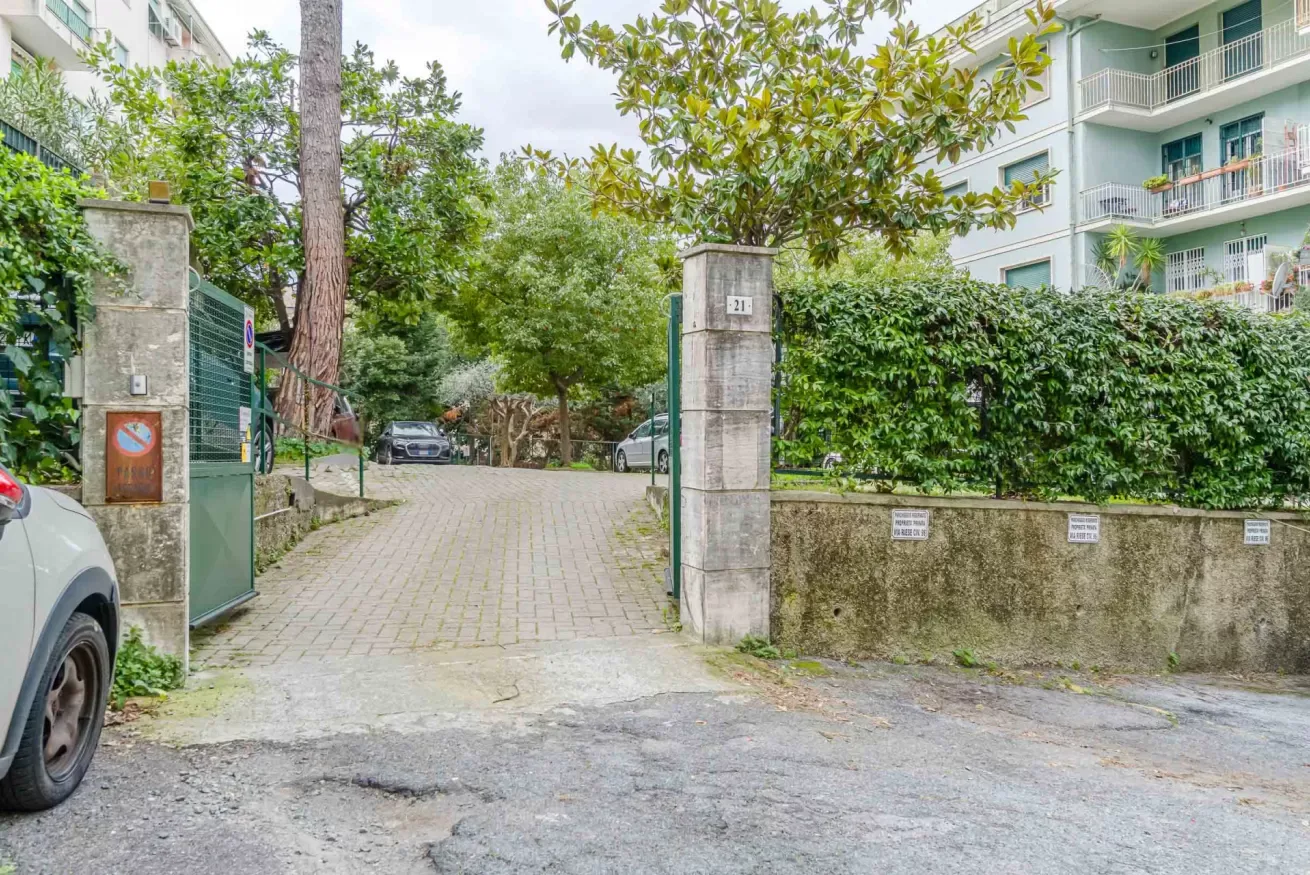 A paved driveway entrance between concrete pillars, with trees and apartment buildings nearby.