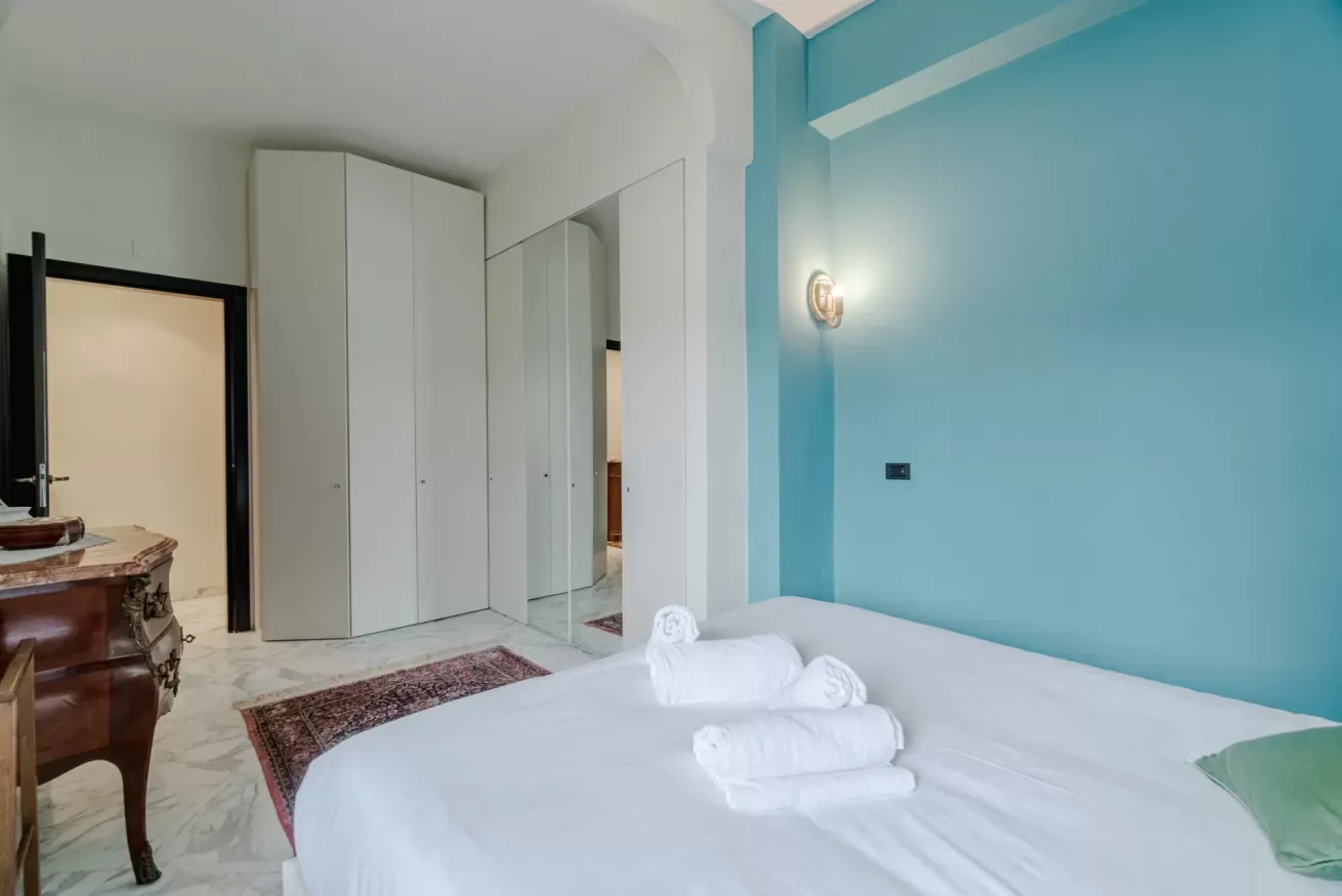A bedroom with a white bed, folded towels, and a turquoise accent wall.