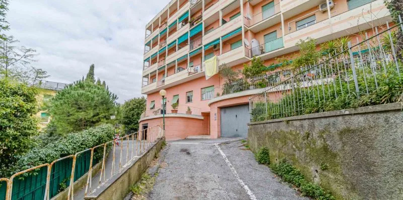 An apartment building entrance opens onto a sloped paved driveway bordered by greenery.