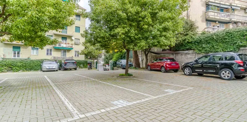 A paved parking area sits beneath leafy trees beside apartment buildings.