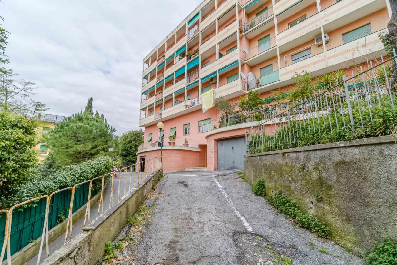 A multistory apartment building stands above a sloped driveway bordered by greenery.