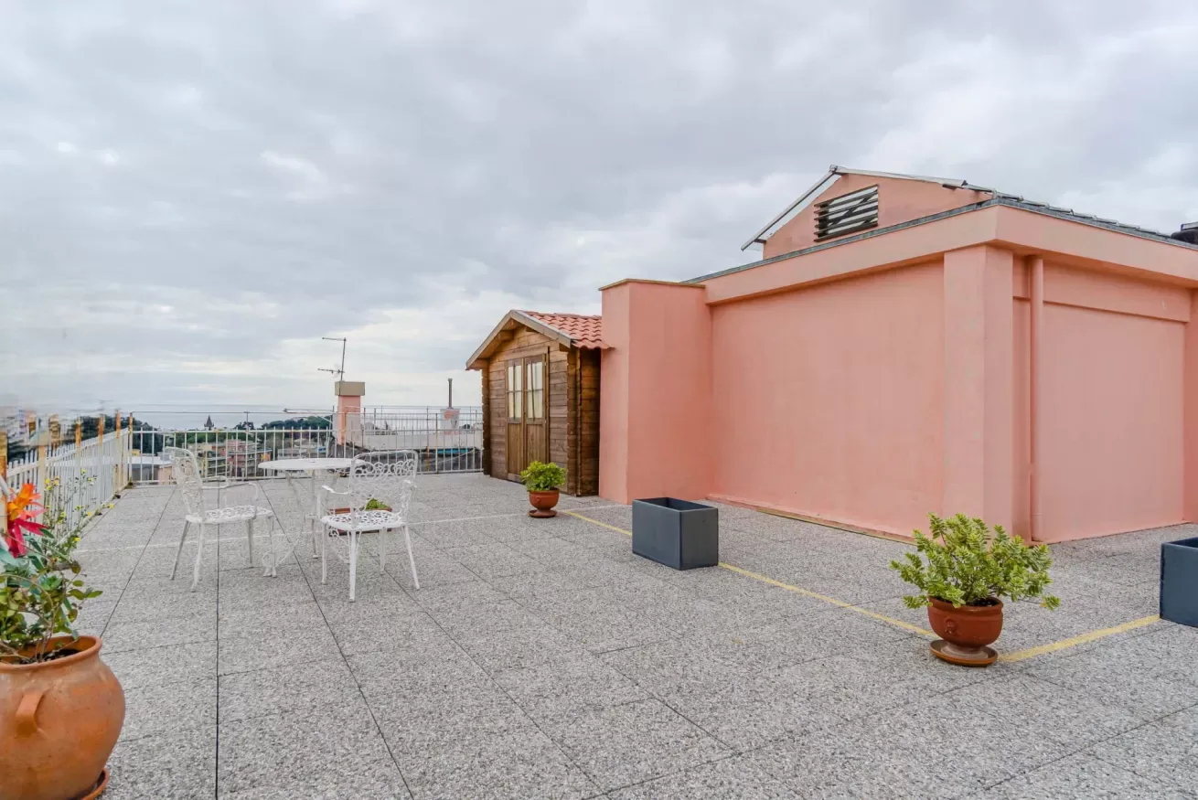 A rooftop terrace with outdoor seating and potted plants beside a pink building.