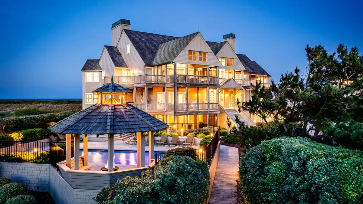 A large illuminated coastal house stands beside a garden path and covered seating area.