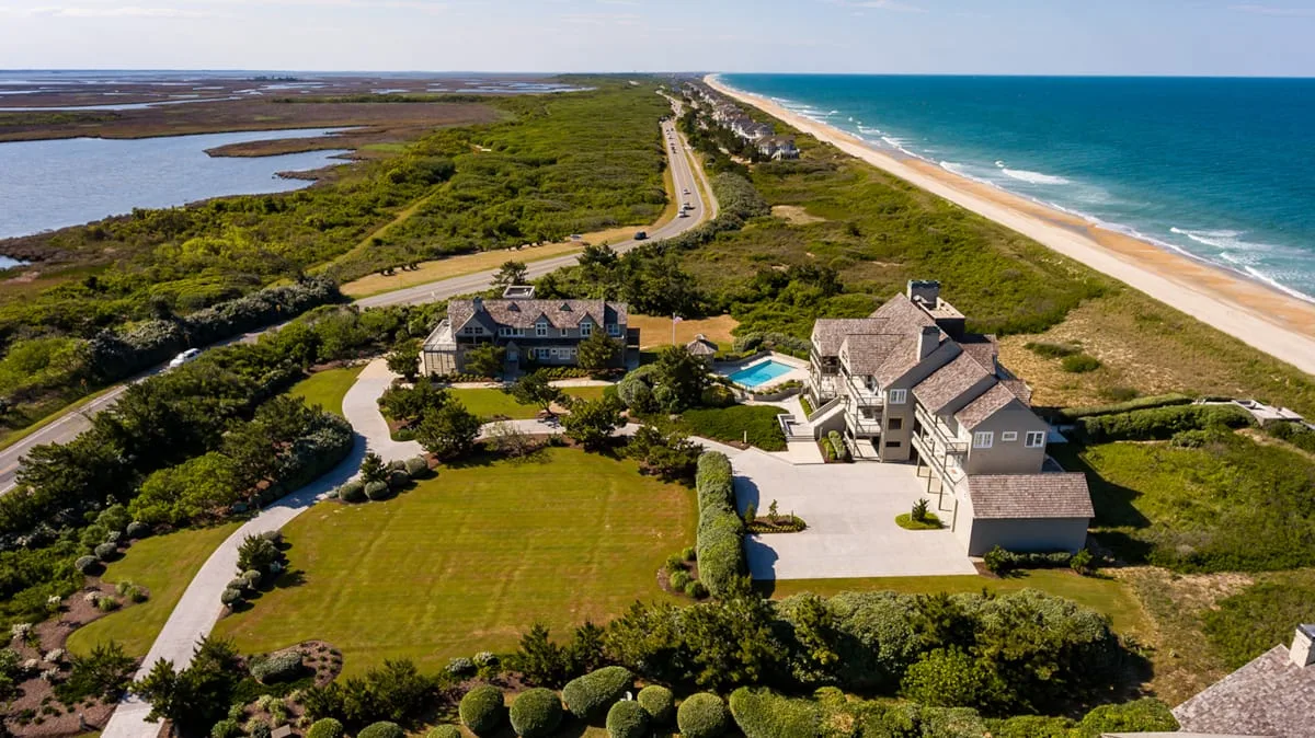 Aerial view of coastal homes beside a beach, road, and green marshland.