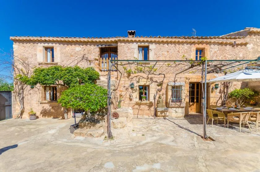 Una casa de piedra da a un patio soleado con una pérgola cubierta de enredaderas.