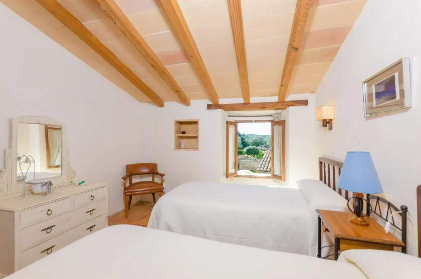 Un dormitorio luminoso con ropa de cama blanca, vigas vistas y una ventana con vistas a la vegetación.
