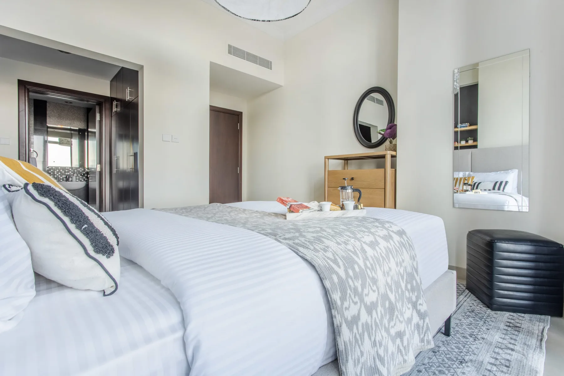 A bedroom with a neatly made bed, patterned pillows, and a round wall mirror.
