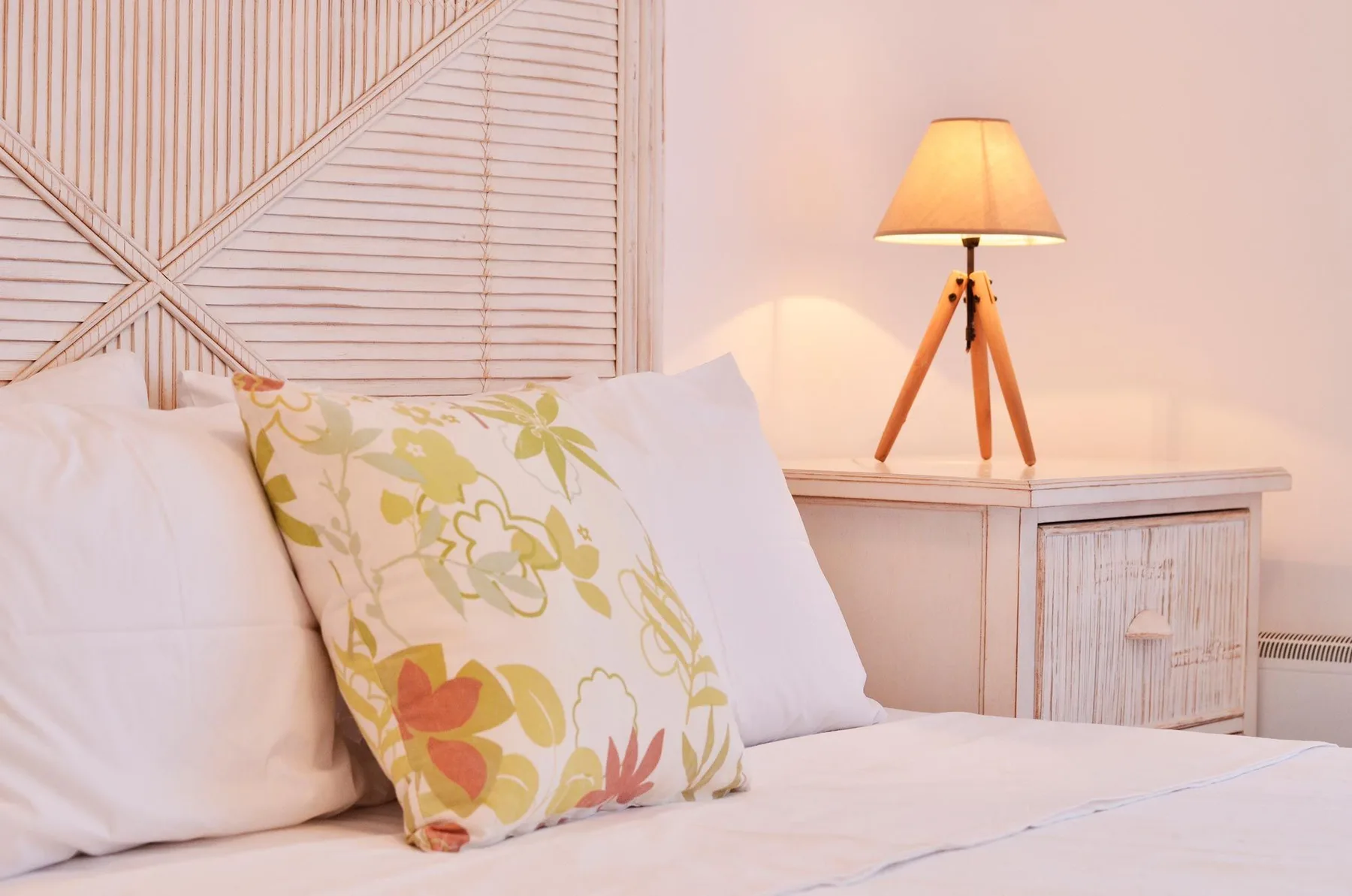 A bedroom with white bedding, a patterned pillow, and a lit bedside lamp.