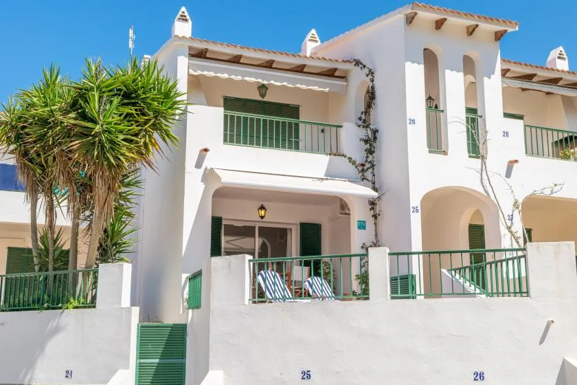 Un edificio blanco de estuco con balcones y barandales verdes se alza bajo un cielo azul despejado.