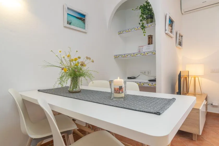 Un comedor luminoso con una mesa blanca, asientos y un jarrón con flores.