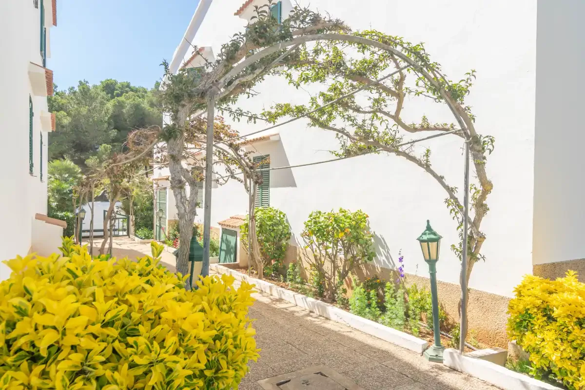 Un camino pavimentado atraviesa un jardín junto a edificios blancos, arbustos y un arco frondoso.