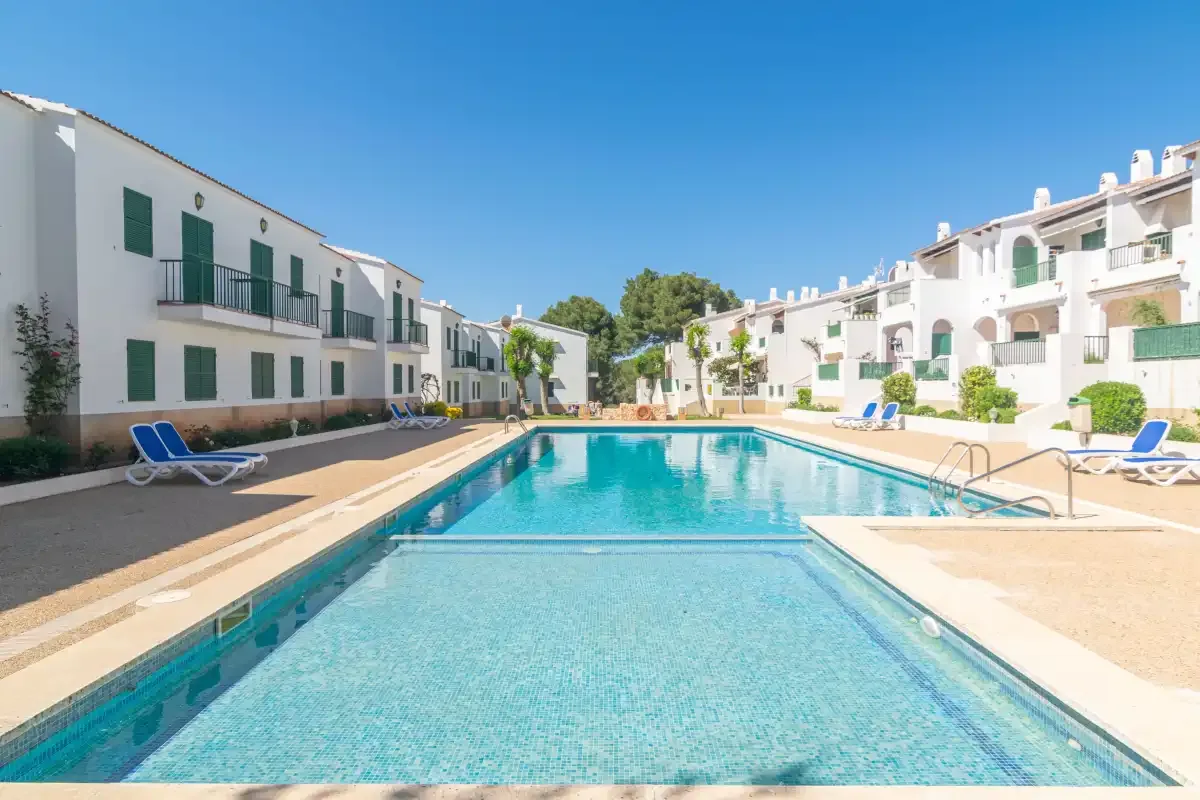 Una piscina exterior se encuentra entre edificios de apartamentos blancos bajo un cielo azul despejado.