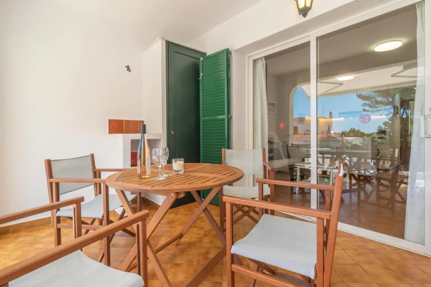 Zona de comedor con suelo de baldosas, mesa redonda de madera y sillas junto a puertas de cristal.